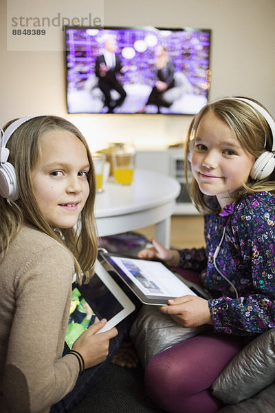 Porträt von lächelnden Schwestern beim Musikhören auf digitalen Tabletts im Wohnzimmer