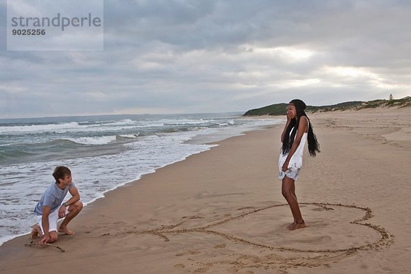 Junger Mann auf einem Knie am Strand  junge Frau in Herzform stehend
