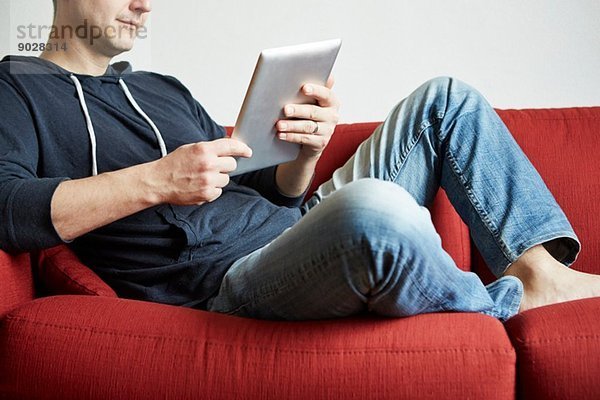Mittlerer Erwachsener Mann  der sich auf dem Sofa entspannt und auf ein digitales Tablett schaut.