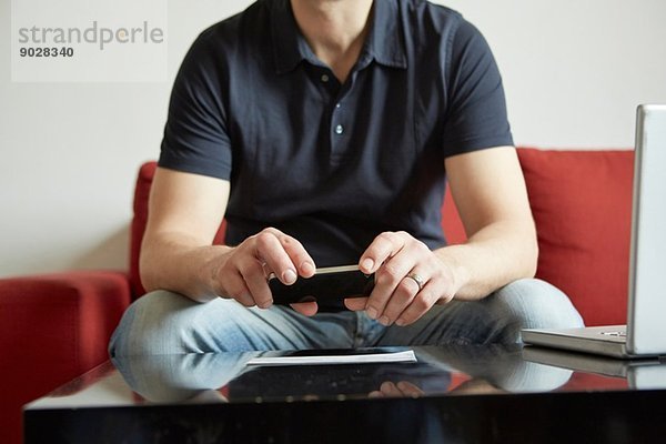 Mittlerer Erwachsener Mann hält Smartphone mit digitalem Tablett und Laptop auf Tisch