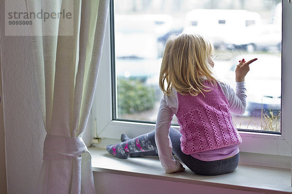 Kleines Mädchen sitzt auf der Fensterbank und schaut aus dem Fenster.