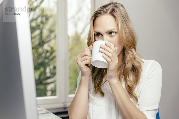 Blonde junge Frau am Schreibtisch  die eine Tasse Kaffee trinkt.