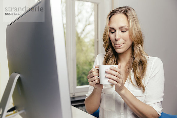 Blonde junge Frau am Schreibtisch  die eine Tasse Kaffee trinkt.