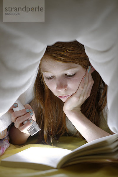 Porträt eines Mädchens  das heimlich ein Buch im Bett unter der Decke liest.