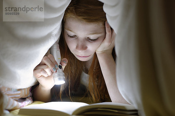 Porträt eines Mädchens  das heimlich ein Buch im Bett unter der Decke liest.