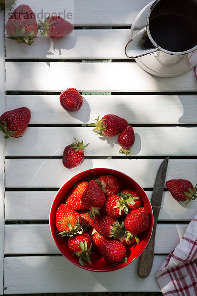 Schale mit Erdbeeren  Messer und Glas auf weißem Holz  erhöhte Ansicht