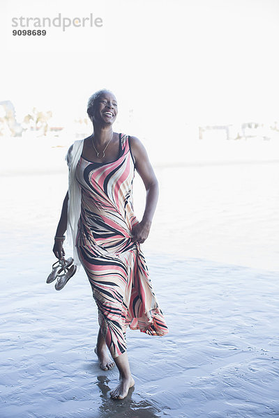 Fröhliche Frau im Kleid  die barfuß am Strand spazieren geht.