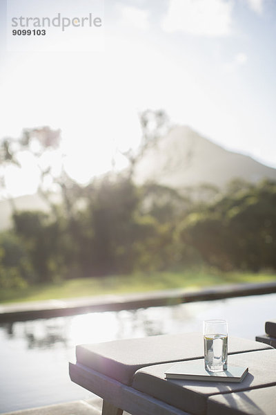 Wasserglas und Buch auf Liegestuhl am sonnigen Poolrand