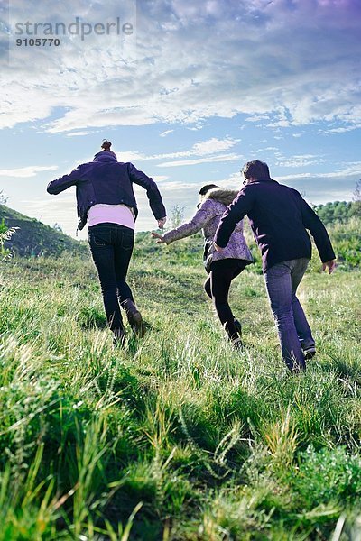 Drei junge erwachsene Freunde rennen den grasbewachsenen Hügel hinauf.