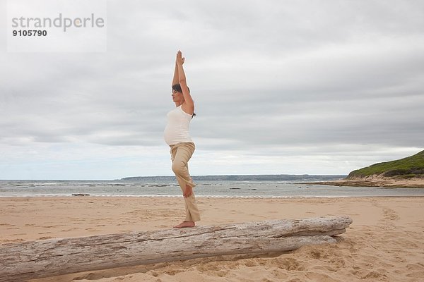 Schwangere mittlere erwachsene Frau beim Yoga am Baumstamm am Strand