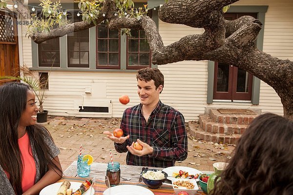 Freunde sitzen am Tisch und teilen sich Grillgut  Mann jongliert mit Früchten