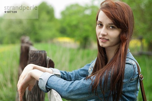Porträt einer jungen Frau  die sich auf einen rustikalen Zaun stützt.