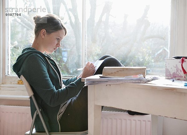 Sechzehnjähriges Mädchen beim Lernen und Schreiben von Notizen neben dem Fenster