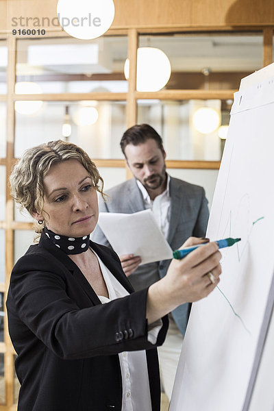 Reife Geschäftsfrau beim Schreiben auf Flipchart mit männlichem Kollegen im Hintergrund im Büro