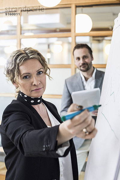 Porträt einer reifen Geschäftsfrau auf Flipchart mit männlichem Kollegen im Hintergrund im Büro