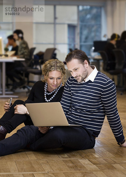 Intelligent gekleidete Geschäftsleute  die den Laptop benutzen  während sie im Büro auf dem Boden sitzen.