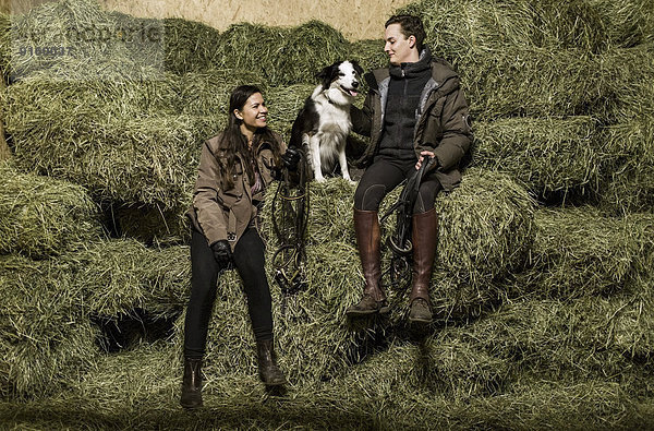 Durchgehende Rückansicht des glücklichen jungen Paares mit Hund auf dem Heuhaufen im Stall