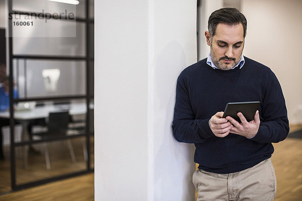 Mittlerer erwachsener Geschäftsmann  der ein digitales Tablett benutzt  während er sich auf eine Säule im Büro stützt.