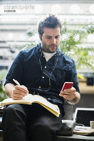 Mittlerer erwachsener Geschäftsmann mit Handy beim Schreiben in einem Buch im Cafe