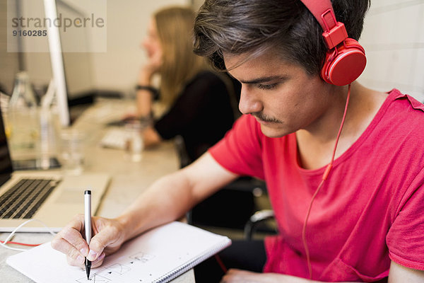 Junger Geschäftsmann mit Kopfhörer beim Schreiben auf Buch mit Kollegen im Hintergrund im Kreativbüro