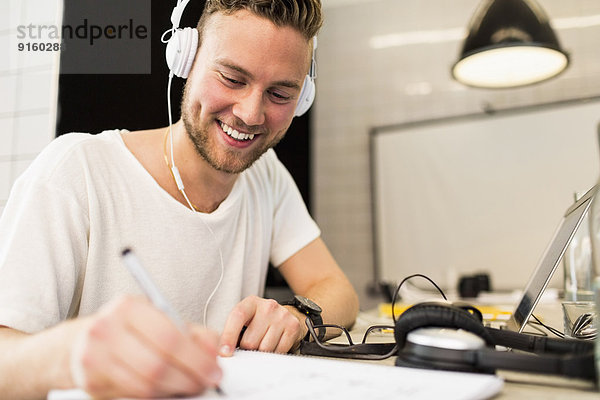 Glücklicher junger Geschäftsmann mit Kopfhörer beim Schreiben im Kreativbüro