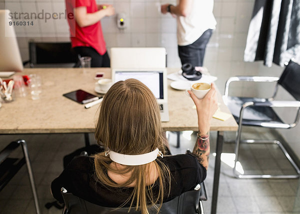 Rückansicht der jungen Geschäftsfrau beim Kaffee mit männlichen Kollegen im Hintergrund im neuen Büro