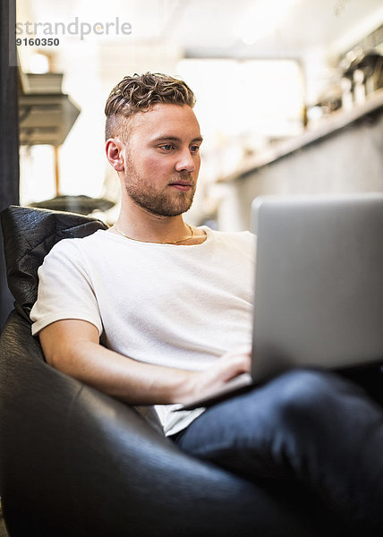 Junger Geschäftsmann mit Laptop  während er im neuen Büro auf einer Bohnentüte sitzt.