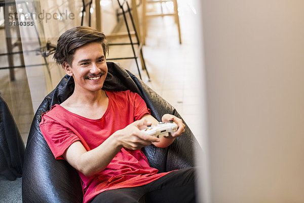 Glücklicher junger Mann spielt Videospiel  während er zu Hause auf einer Bohnentüte sitzt.