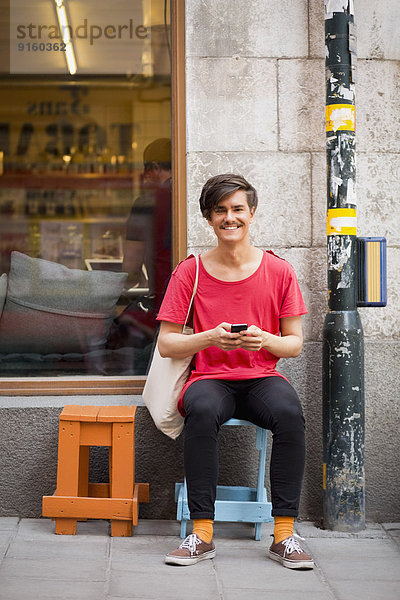 Ganzflächiges Porträt eines glücklichen jungen Mannes  der auf einem Handy auf dem Bürgersteig SMS schreibt