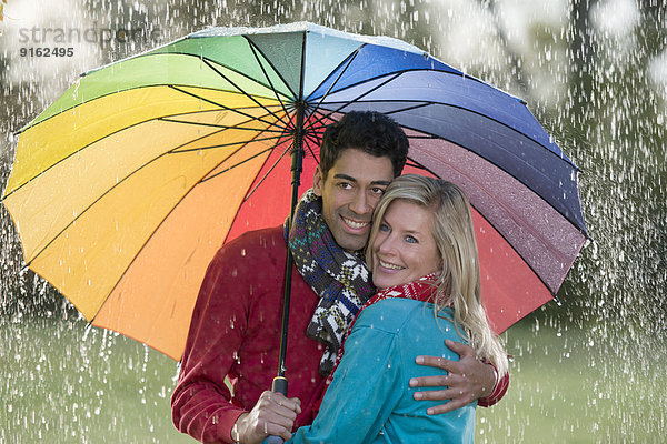 Lächelndes Paar unter regenbogenfarbenem Schirm genießt den Regen