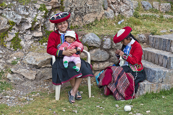 Zwei Frauen der Quechua-Indianer in traditioneller Kleidung  eine lächelnde Frau sitzt auf einem Stuhl und hält ein Kleinkind  eine zweite Frau sitzt auf einer Steintreppe und strickt  Cinchero  Urubambatal  Peru