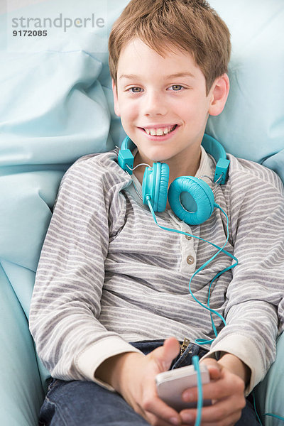 Portrait des lächelnden Jungen mit Smartphone und Kopfhörer auf Sitzsack liegend