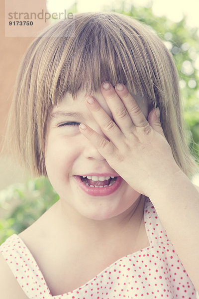 Porträt eines lachenden kleinen Mädchens  das ein Auge mit der Hand bedeckt.