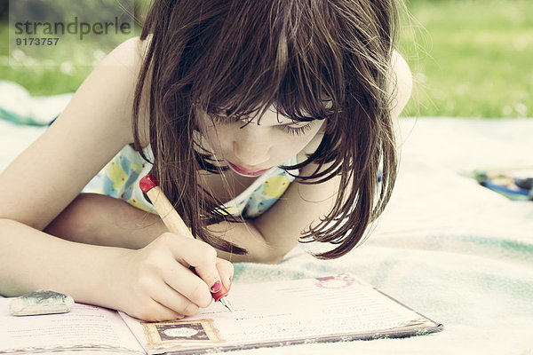 Porträt eines kleinen Mädchens  das auf einer Decke im Garten liegt und ein Tagebuch schreibt.