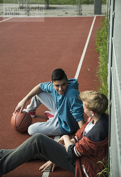 Zwei Jungen mit Basketball  die sich gegen den Zaun lehnen.