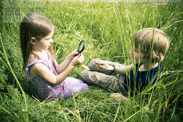 Bruder und Schwester sitzen auf der Wiese und spielen mit der Lupe.