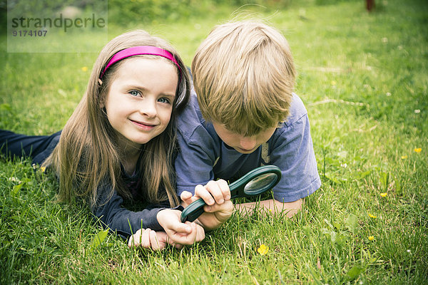 Bruder und Schwester liegen auf der Wiese und beobachten die Blume mit Lupe.