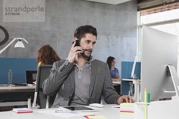 Porträt eines Mannes  der mit seinem Smartphone an seinem Arbeitsplatz im Büro telefoniert.