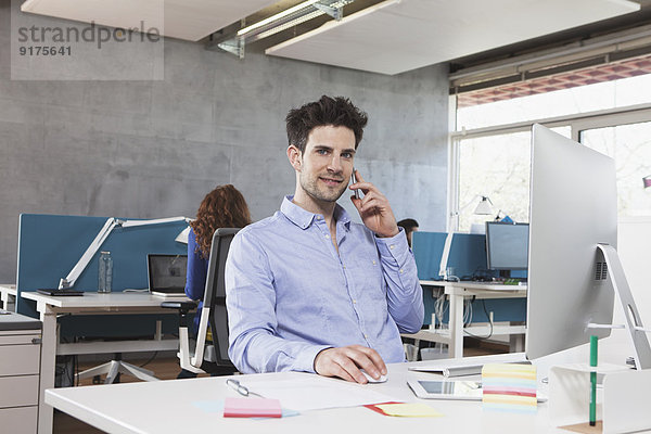 Porträt eines Mannes  der mit seinem Smartphone an seinem Arbeitsplatz im Büro telefoniert.