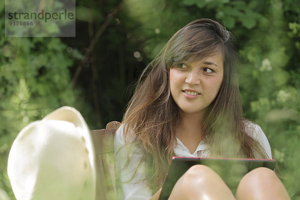 Porträt einer jungen Frau mit digitalem Tablett auf einer Holzbank im Garten