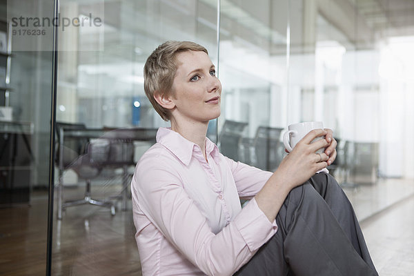 Deutschland  München  Geschäftsfrau im Büro  sitzend auf dem Boden mit Kaffeetasse