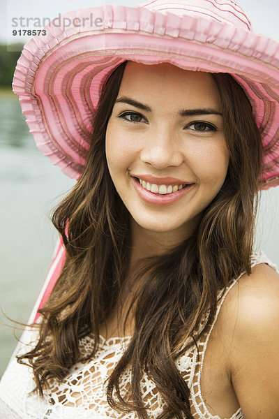 Porträt einer lächelnden jungen Frau am Strand mit rosa Sommerhut
