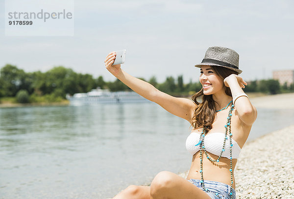 Porträt einer jungen Frau  die einen Selfie mit ihrem Smartphone am Strand nimmt.