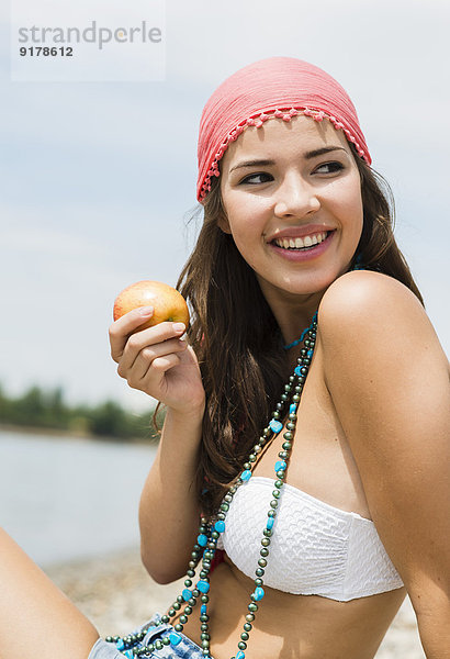 Porträt einer lächelnden jungen Frau mit Apfel am Strand