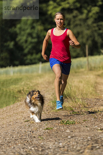 Junge Frau beim Joggen mit Hund auf dem Feldweg