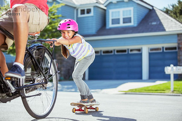 Vater auf Fahrrad zieht Tochter auf Skateboard