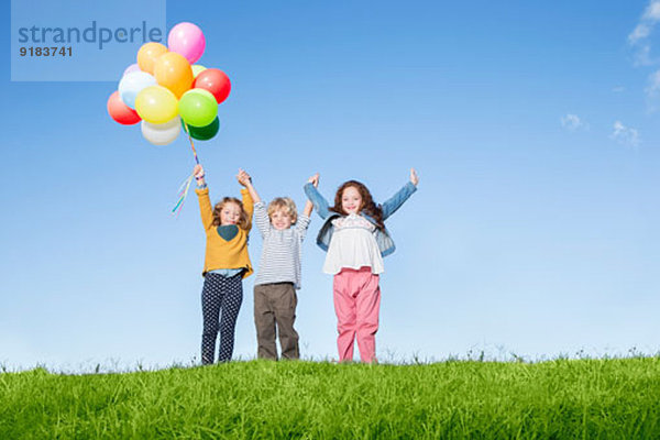 Kinder mit Luftballons jubelnd auf dem grasbewachsenen Hügel