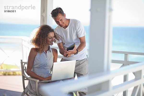 Paar mit Blick auf digitales Tablett und Laptop auf dem Balkon des Strandhauses