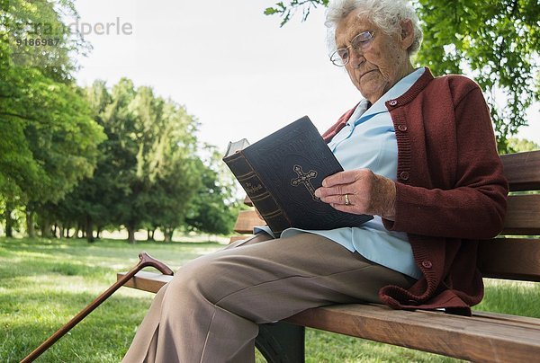 Seniorin sitzt auf der Parkbank und liest die Bibel.