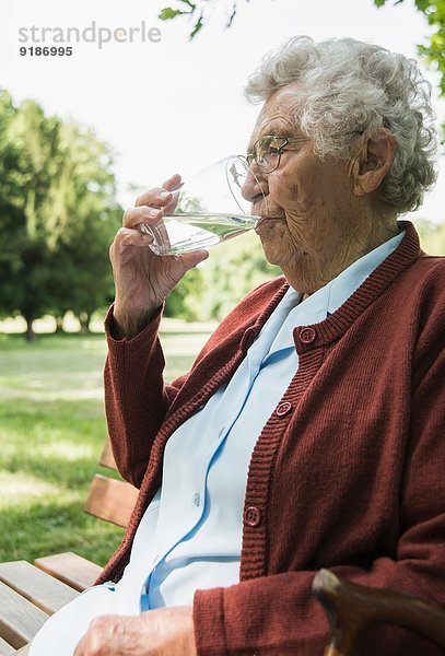 Seniorin sitzt auf einer Parkbank und trinkt ein Glas Wasser.